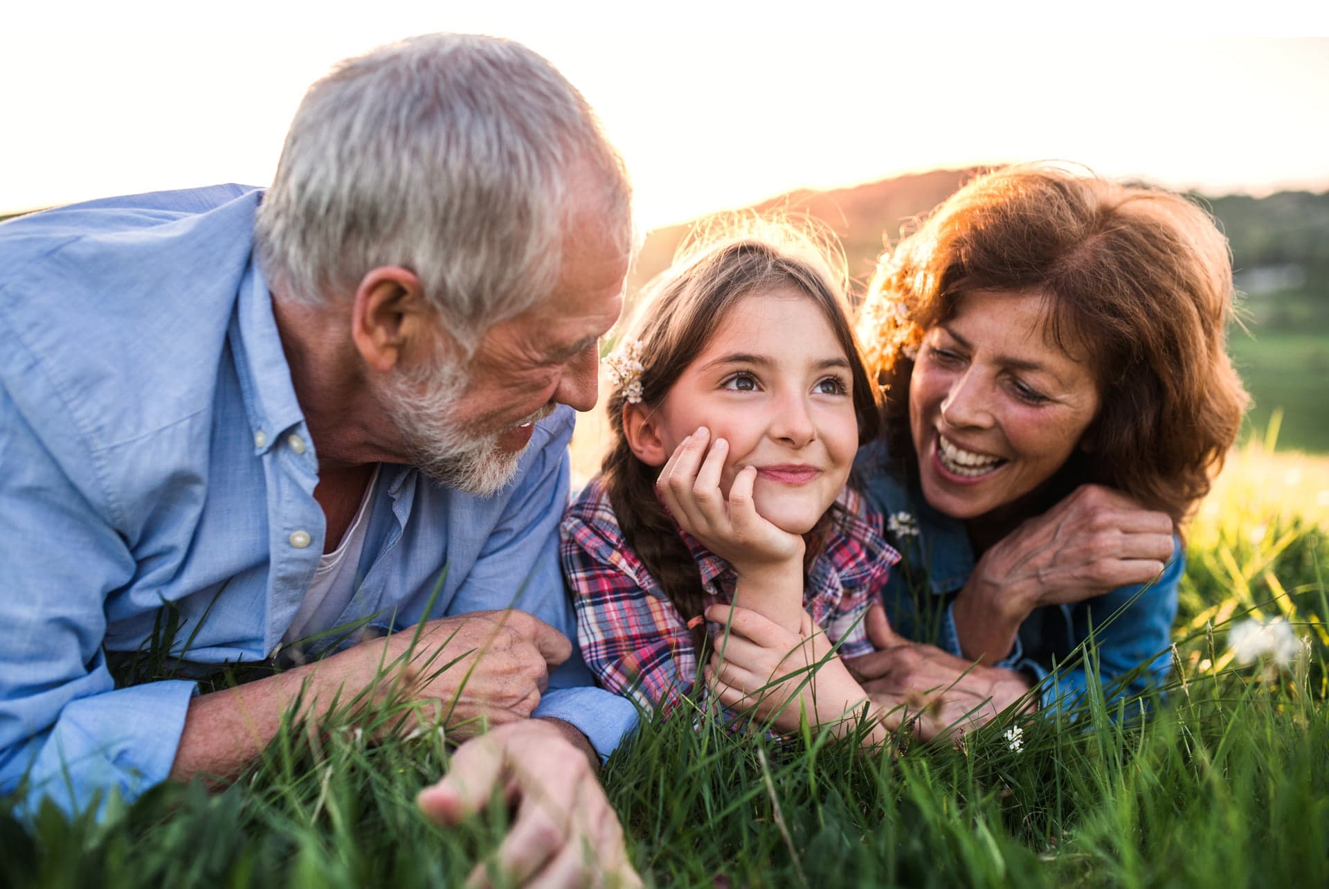Un abuelo y una abuela conversan y ríen con su nieta, recostados sobre la grama al atardecer.